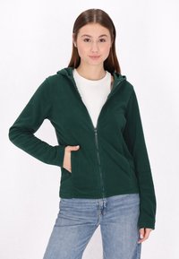 Felpa con zip di colore verde scuro, dotata di cappuccio, tasche frontali, maniche lunghe e una texture morbida; abbinata a jeans azzurri chiari.