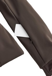 Hnedá tkanina s hladkou textúrou obsahuje skrytú kapsu, v ktorej sa nachádza biela karta. Dizajn zahŕňa čisté línie a priliehavý strih.