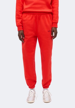 Person iført lyse røde joggingbukser med elastiske manchetter og hvide sneakers, stående mod en ensfarvet lys baggrund.