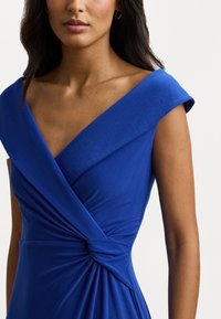 Blue formal dress with a deep V-neck, cap sleeves, and a twisted detail at the waist, made from a smooth, matte fabric.