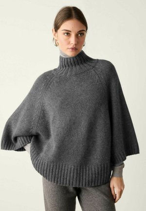 Frau trägt einen lockeren grauen Rollkragen-Strickpullover mit weiten Ärmeln und passenden grauen Hosen und steht vor einem schlichten Hintergrund.