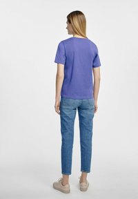 Fioletowa koszulka z krótkim rękawem, o okrągłym dekolcie, w połączeniu z niebieskimi dżinsami. Model w beżowych sneakersach. Gładka tekstura materiału.