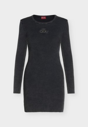 Femme en robe noire cintrée à manches longues, tenant un petit sac à main métallique, avec des bottes noires montantes, sur un fond clair uni.