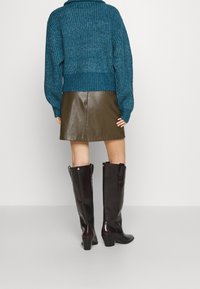 Pull en tricot teal avec ourlet côtelé, associé à une jupe en cuir marron et des bottes à talons noires hautes, vu de dos.