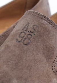 Gros plan sur une chaussure en daim marron montrant le logo AS 98 embossé et des coutures détaillées près de l'ouverture.