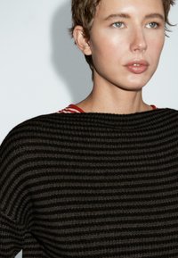 Osoba z krótkimi jasnobrązowymi włosami i niebieskimi oczami, nosząca czarny sweter o teksturowanym splotach na czerwono-białą w paski koszulę.