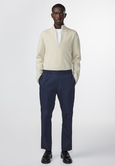 Crème gebreide half-zip trui gecombineerd met marineblauwe elastische taillebroek. Het model draagt zwarte schoenen en toont soepele texturen en nauwsluitende vormen.