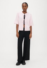 Woman wearing a light pink short-sleeve cardigan tied at the front, black cropped top, wide-leg black pants, and black platform shoes.