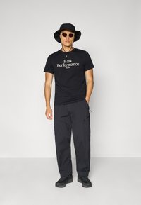 Sort t-shirt med "Peak Performance" logo, parret med sorte cargo-bukser, sort bøttehat og solbriller. Fodtøjet er sorte vandrestøvler.