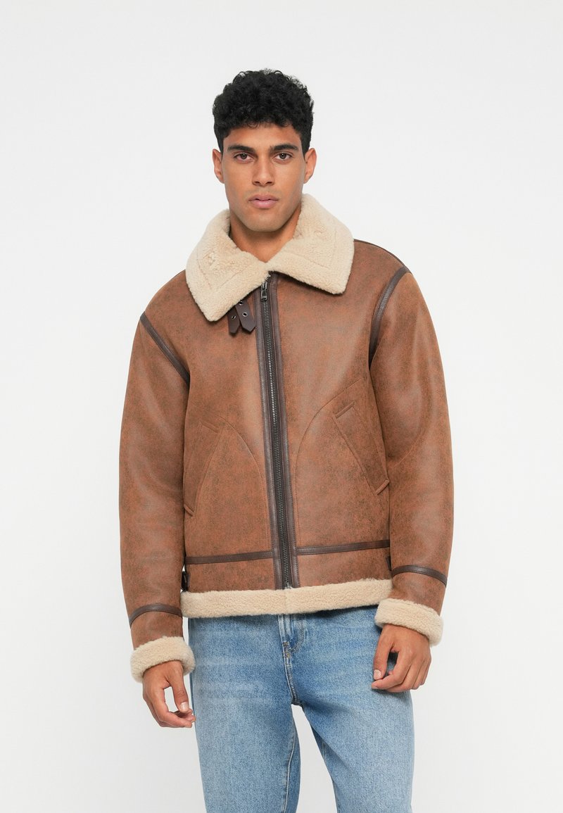 Uomo che indossa una giacca in shearling marrone con colletto e polsini in pelliccia beige, abbinata a jeans azzurri e in piedi contro uno sfondo bianco.