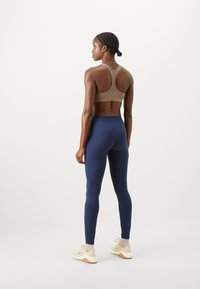 Beige sports-bh med racerback-design, kombinert med marineblå høytaljede tights. Modellen har på seg lyse joggesko med teksturert såle.