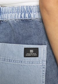 Světle modré denimové kalhoty s elastickým pasem, na kapse mají černou nášivku se nápisem "BDG STYLE NO. 003/47 DESIGNED TO ADAPT."