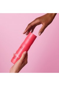 Roze fles Wella Color Brilliance shampoo, gladde textuur, lichte kromming, vastgehouden door een hand, met een andere hand die naar de dop reikt.