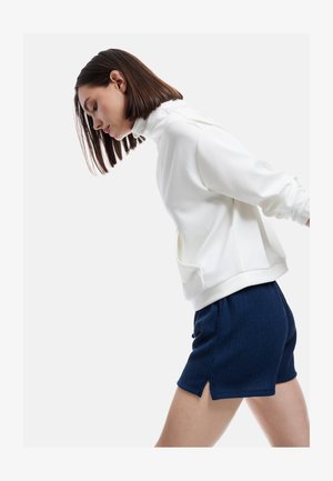 Ung kvinde med glat mørkt hår iført en hvid højhalset sweater og marineblå teksturerede shorts, der læner sig let fremad.