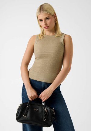 Blondynka w złotych kolczykach kołach i beżowej bluzce bez rękawów trzyma czarną torebkę Guess, stojąc na białym tle.