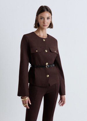 Frau mit kurzen braunen Haaren, die eine dunkelbraune Jacke mit goldenen Knöpfen, schwarzen Gürtel, passende Hosen und goldenen Schmuck trägt, stehend.