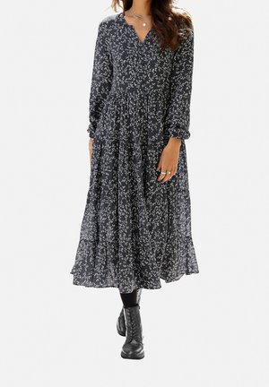 Czarna sukienka w kwiaty z długimi rękawami i dekoltem w serek, z warstwową spódnicą i zwiewną sylwetką. Noszona z czarnymi botkami za kostkę.