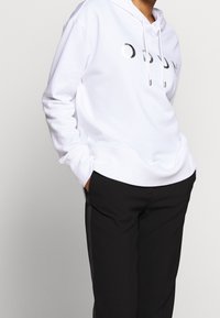 Sudadera blanca con un logotipo frontal en fuente metálica, con capucha ajustable, corte relajado y bolsillos laterales, combinada con pantalones negros.
