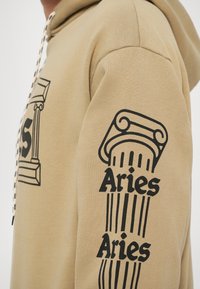 Bež hudica s grafikom klasičnog stuba i riječi "Aries" otisnutom u crnoj boji na rukavu i prednjem dijelu. Tekstuirana tkanina.