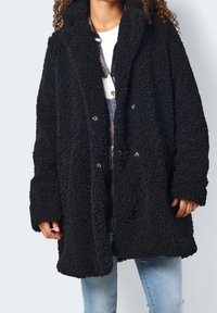 Svart teddyjacka med en texturerad, fluffig tygkvalitet. Har krage, knappstängning och vida ärmar, bärs över en vit skjorta.