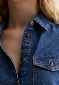 Närbild av en person i en blå denimskjorta med synliga knappar, krage och bröstficka, som visar en del av deras nacke och hår.