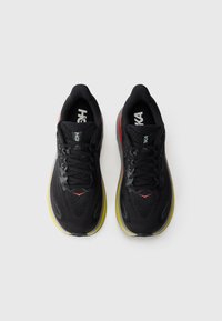 Pantofi sport negri cu un superior din plasă, talpă galbenă, accente roșii și gri, și șireturi negre. Prezintă un design aerodinamic și ușor.