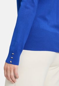 Kék kötött pulóver bordás mandzsettákkal, a bal ujján három arany színű gombbal. Simább textúra, testhezálló fazon.