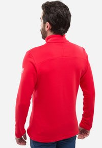 Veste mi-saison en polaire rouge avec un col haut et des manches longues. Texture lisse et design ajusté avec des détails de couture discrets.