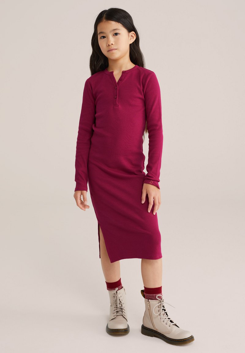 Abito fucsia a maniche lunghe con scollatura henley e spacco laterale, abbinato a stivaletti beige con lacci e calze a righe rosse.