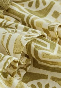 Tissu doux avec des motifs géométriques et organiques abstraits en vert olive sur un fond crème, montrant des plis et des coutures visibles.