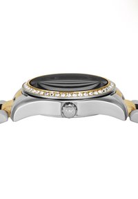 Montre-bracelet argentée et dorée avec un cadran noir rond, une lunette en cristal et un bracelet métallique. Présente un logo MK bien en vue sur la couronne.