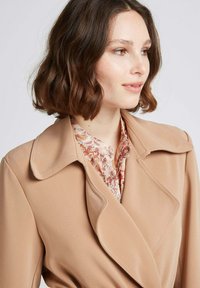 Blazer beige avec un large col et un design ajusté, superposé à un chemisier floral léger présentant des nuances de rose et d'orange. Texture lisse.