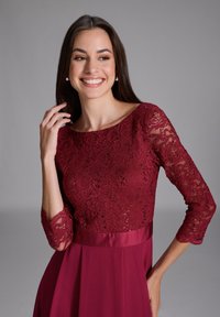 Burgundy spetsklänning med trekvartsärmar, med en texturerad kropp och en slät satinsmidja, flödande ut i en lös kjol.