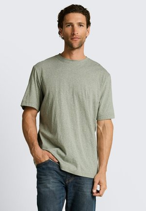 Mann mit kurzen, lockigen Haaren, der ein hellgraues T-Shirt mit kurzen Ärmeln und eine dunkelblaue Jeans trägt, vor einem schlichten Hintergrund steht.