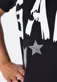 Camiseta de algodón negra con grandes letras blancas y un acento de estrella plateada texturizada, destacando un diseño casual y una textura suave.