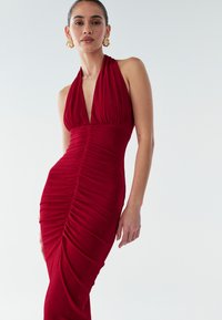 Moteris, vilkinti be rankovių tamsiai raudoną susiraukinę halter stiliaus suknelę ir auksines suktas auskarų kilpas, prieš paprastą šviesią fono.