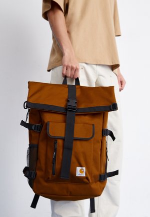 Personne tenant un grand sac à dos couleur rouille avec des bretelles noires et une poche avant, portant un pantalon beige et une chemise brun clair.