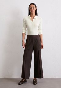 Rippiertes, cremefarbenes Langarm-Polo-Shirt kombiniert mit weiten, dunkelbraunen Hosen. Das Modell trägt dunkle Slipper. Schlichte, strukturierte Silhouette.