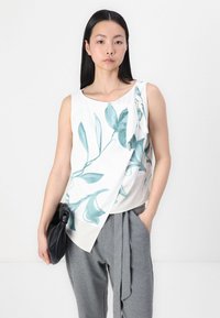 Haut sans manches en tissu blanc avec des imprimés botaniques verts, doté d'un détail noeud à l'épaule gauche et d'un ourlet asymétrique.
