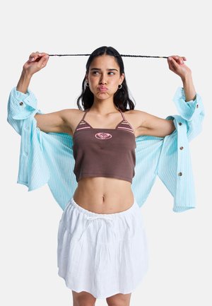 Femme tirant ses longs cheveux tressés sur le côté avec les deux mains, portant un crop top marron, une jupe blanche et une chemise à rayures bleu clair.