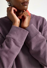 Felpa viola con polsini a coste, indossata sopra una camicia rossa con colletto. In primo piano, mani che regolano il colletto, mettendo in evidenza la trama del tessuto.