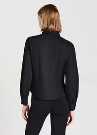 Schwarze Bluse mit Kragen und langen Ärmeln, verfügt über eine taillierte Passform und glattes Material. Die Rückansicht zeigt einen leicht abgerundeten Saum und minimale Details.