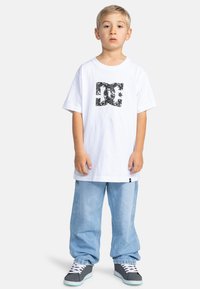 Bílý bavlněný tričko s černým grafick�ým logem, spárované se světle modrými volnými džínami a šedými teniskami s bílými tkaničkami a zelenými akcenty.