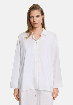 Mujer con cabello castaño claro vistiendo una camisa blanca de botones holgada y pantalones blancos a juego frente a un fondo liso.