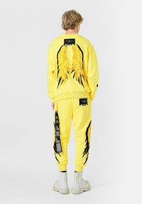 Sudadera y pantalones de algodón amarillos con grandes gráficos de alas en la parte posterior y los lados, complementados por acentos y etiquetas negras.
