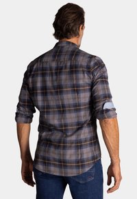 Geruite shirt in marineblauw, grijs en bruin met opgerolde mouwen. Kenmerkt zich door een gebogen zoom en contrasterende stofdetails bij de mouwmanchetten.