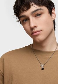 Felpa marrone con colletto rotondo e maniche corte; collana in argento con ciondolo circolare e una perla nera.