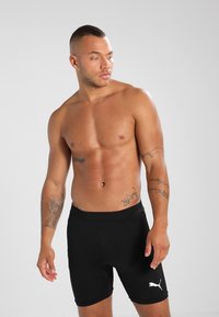 Svarte treningsshorts laget av stretchy stoff, med en tettsittende passform og hvit logo på siden. Modellen har skjegg og viser ingen overkropp.