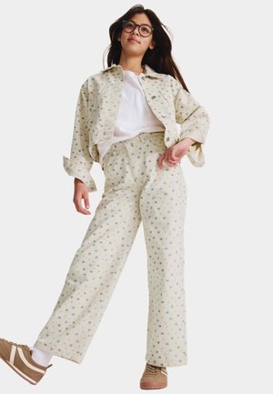 Joven con cabello largo y gafas, que lleva una chaqueta crema con flores y pantalones anchos a juego, camisa blanca y zapatillas beige.