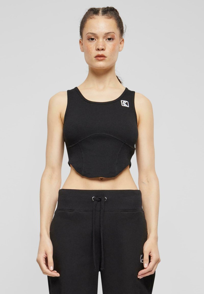 Must mustriba crop-top logodegusega, laiad rihmad ja tihe disain, koos mustade tõmblukkudega spordipükstega.